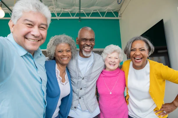 A group of older adults from diverse backgrounds standing close together indoors, smiling and laughing with their arms around each other.