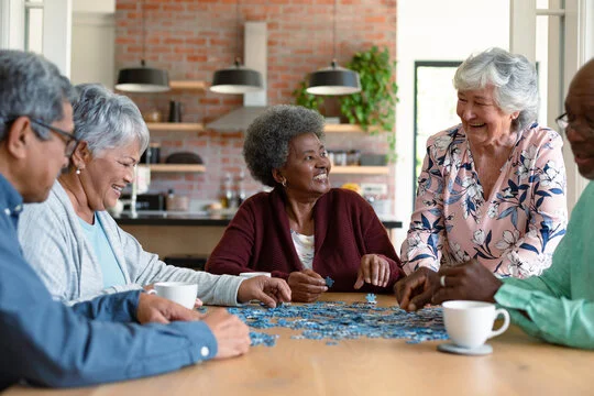 A diverse group of older adults sitting around a table indoors, smiling and working together on a jigsaw puzzle while drinking coffee.