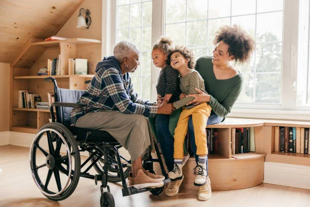 An older adult in a wheelchair sitting by a window with two young children and a woman, smiling and sharing a warm family moment in a cozy room.